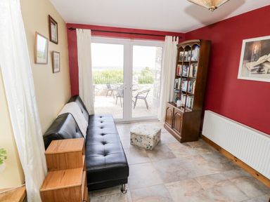 A sitting room with a black leather sofa a wooden bookshelf a small ottoman and patio doors leading outside at Prague House in Lettermore County Galway