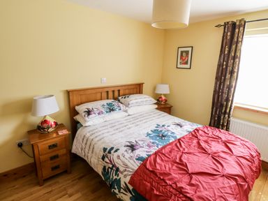 A bedroom with a wooden bed and floral bedding with a red throw a wooden nightstand with a lamp on each side a window with floral curtains and framed picture on the wall at Prague House in Lettermore County Galway