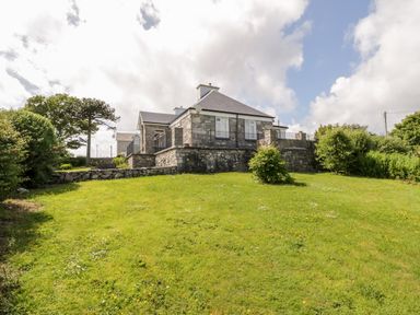 A stone house with a slate roof on a grassy lawn with bushes and trees at Prague House in Lettermore County Galway