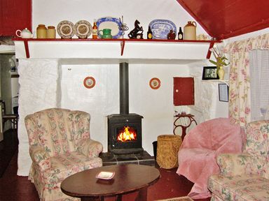 A living room with a wood stove fireplace two floral armchairs a round wooden table and shelves with decorative plates at Carthy's Cottage Kilbrien near Dungarvan County Waterford