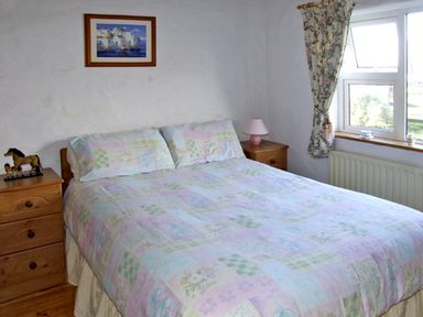 A bedroom with a bed between two wooden bedside tables with lamps and a window with floral curtains at Delia's Cottage in Ballinrobe County Mayo
