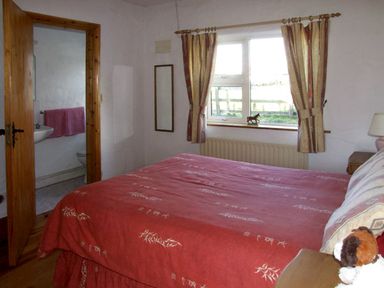A bedroom with a red bedspread a window with curtains a wooden door leading to a bathroom at Delia's Cottage in Ballinrobe County Mayo