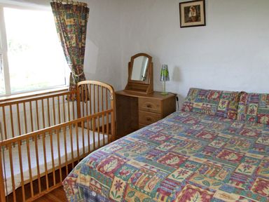 A bedroom with a double bed a baby cot a wooden dresser with a mirror and a lamp and a window with patterned curtains at Delia's Cottage in Ballinrobe County Mayo
