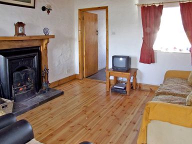 A living room with a fireplace a wooden floor a small table with a TV and a sofa with curtains at Delia's Cottage in Ballinrobe County Mayo