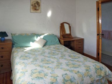 A bedroom with a bed covered in floral bedding two green pillows a bedside table with a lamp a dressing table with a mirror and an open door leading to a bathroom at Delia's Cottage in Ballinrobe County Mayo