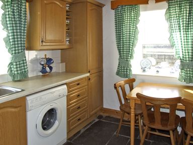 A kitchen with wooden cabinets a washing machine a wooden table and chairs with green check curtains at Delia's Cottage in Ballinrobe County Mayo