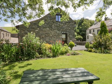 A garden with a stone house surrounded by flowers and a green picnic table at Woodside Barn in Pennington near Ulverston