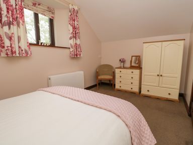 A bedroom with a bed a window with floral curtains a chair a dresser with a mirror and flowers and a wardrobe at Woodside Barn in Pennington near Ulverston