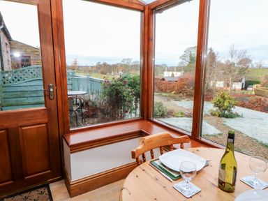 A dining area with a round wooden table set with plates glasses and a wine bottle near large windows overlooking a garden at The Stable in Milton near Brampton