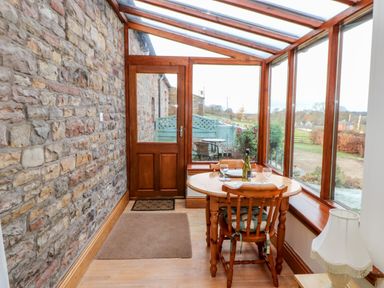 A small dining area with a round wooden table and two chairs next to a glass door and windows with a stone wall at The Stable in Milton near Brampton