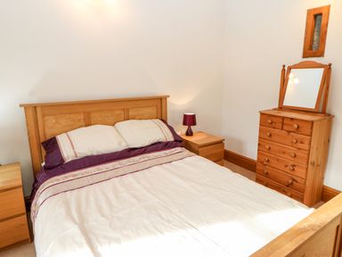 A bedroom with a wooden bed frame and headboard a large bed with cream and purple bedding two wooden bedside tables one with a small purple lamp a wooden dresser with a mirror on top and a small rectangular window on the wall at The Stable Milton near Brampton