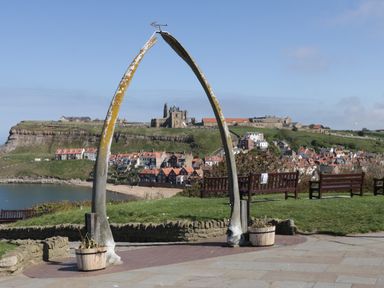 A coastal village with red roof houses and a historic abbey viewed through a large whale bone arch with benches on green grass near the shore at Barn Cottage in High Normanby near Robin Hood's Bay