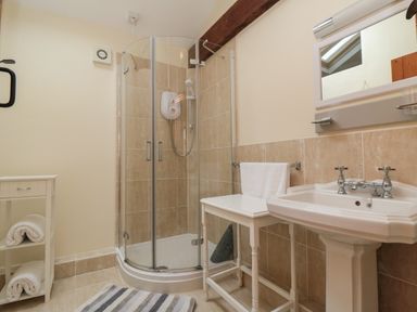 A bathroom with a corner shower, a pedestal sink, a small table with a towel, a striped rug, and a small shelving unit with towels at Barn Cottage High Normanby near Robin Hood's Bay