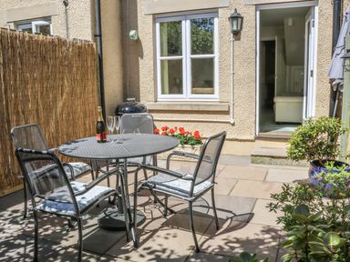 A small outdoor patio with metal table and chairs with cushions and a bottle with glasses at Bramble Cottage in Sedbergh
