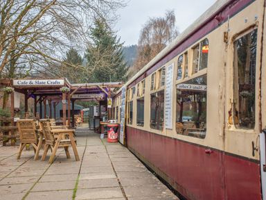 An outdoor seating area with wooden chairs and tables next to a cafe and a vintage train carriage at No 1 Railway Cottages in Betws-y-Coed