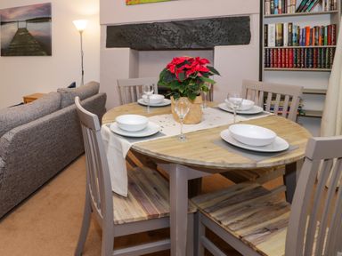 A dining area with a round wooden table set with plates and glasses a red flower centerpiece and wooden chairs next to a grey couch a floor lamp and a bookshelf in No 1 Railway Cottages Betws-y-Coed