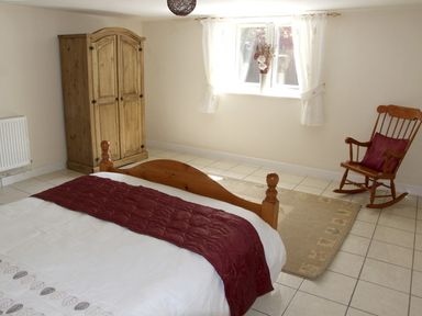 A bedroom with a wooden bed a wardrobe a rocking chair a window with curtains and a rug on tiled floor at Rosewaters Cottage in Astley Burf near Stourport-on-Severn