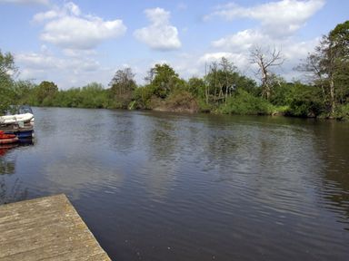 A river with green trees along the bank and a wooden dock with small boats tied up at Rosewaters Cottage in Astley Burf near Stourport-on-Severn