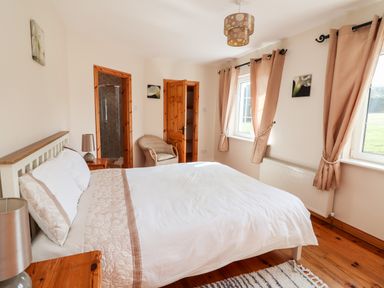 A bedroom with a double bed two windows with curtains a wicker chair and an open door showing a shower at Fern View House Beaufort near Killorglin County Kerry