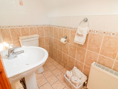 A bathroom with a white sink, toilet, hand towels, a basket with towels, and beige tiled walls and floor at Fern View House in Beaufort near Killorglin County Kerry
