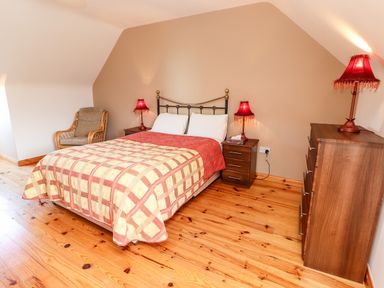 A bedroom with a double bed flanked by two bedside tables with lamps a wooden dresser with a lamp and a chair in the corner at Fern View House in Beaufort near Killorglin County Kerry