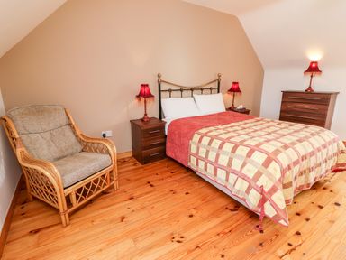 A bedroom with a bed and checkered bedspread two bedside tables with lamps a cushioned wicker chair and a chest of drawers at Fern View House in Beaufort near Killorglin County Kerry