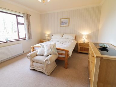 A bedroom with a wooden bed and nightstands with lamps and an armchair with pillows near a window at Brynteg in Llwyngwril