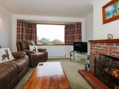 A living room with brown leather chairs a wooden coffee table a brick fireplace and a window with plaid curtains at Brynteg in Llwyngwril