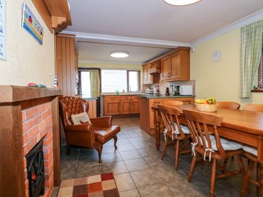 A kitchen and dining area with wooden cabinets and furniture a brown leather armchair a brick fireplace and a table with chairs and fruit at Brynteg in Llwyngwril
