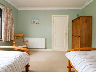 A bedroom with two wooden beds with striped bedding a wooden chair with a cushion a floral curtain a wooden wardrobe and a white door at Brynteg in Llwyngwril