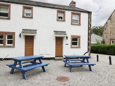 Two blue picnic tables on a gravel area outside a white building with wooden doors and windows at William Court Cottage in Irton Hall near Wastwater