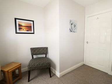 A corner of a room with a patterned chair two stacked wooden stools two pictures on the walls and a closed white door at William Court Cottage in Irton Hall near Wastwater