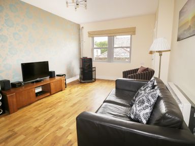 A living room with a black leather sofa, striped armchair, television on wooden stand, wood stove, and wooden floor at William Court Cottage Irton Hall near Wastwater