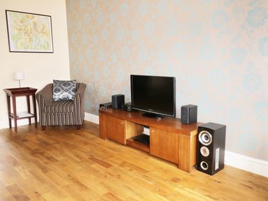 A room with a striped armchair a wooden side table with a lamp a wooden TV stand with a flat screen TV and speakers at William Court Cottage Irton Hall near Wastwater