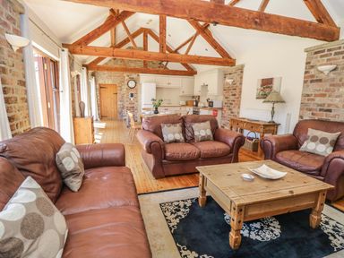 A living area with brown leather sofas and a wooden coffee table opens to a kitchen with wooden beams at Wheelhouse Cottage in Sowerby outskirts of Thirsk
