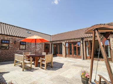 An outdoor patio with wooden chairs and table under an orange umbrella and a wooden swing at Wheelhouse Cottage in Sowerby outskirts of Thirsk