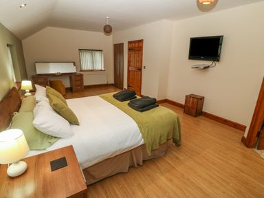 A bedroom with a double bed green bedspread and pillows bedside tables with lamps a dressing table and chair a wall-mounted television wooden doors and wooden floor at Cefn-Yr-Efail in Golan near Porthmadog