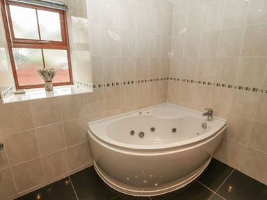 A corner bathtub with jets in a tiled bathroom with a frosted window and a vase of dried flowers at Cefn-Yr-Efail in Golan near Porthmadog