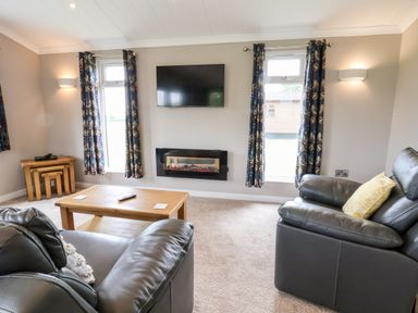 A living room with leather sofas a wooden coffee table a wall-mounted television and curtains at Callow Lodge 15 Beaconsfield Farm Holiday Park near Shrewsbury