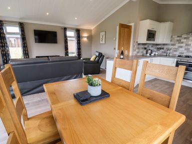 A dining table with chairs next to a kitchen and living room with leather sofas and a wall-mounted tv at Callow Lodge 15 Beaconsfield Farm Holiday Park near Shrewsbury