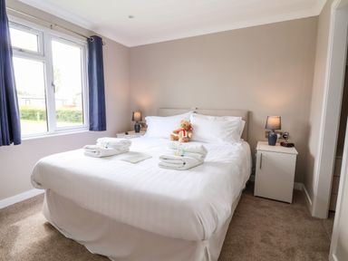 A bedroom with a large bed white bedding towels on bed teddy bear on pillows bedside tables with lamps and window with curtains at Callow Lodge 15 Beaconsfield Farm Holiday Park near Shrewsbury
