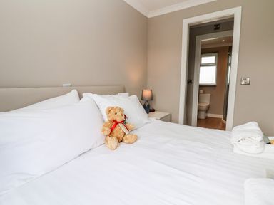A bedroom with a bed featuring white linens and a teddy bear a bedside table with a lamp and a bathroom visible through a doorway at Callow Lodge 15 Beaconsfield Farm Holiday Park near Shrewsbury