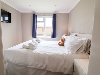 A bedroom with a bed white linens folded towels a teddy bear window with blue curtains and a wall-mounted television at Callow Lodge 15 Beaconsfield Farm Holiday Park near Shrewsbury