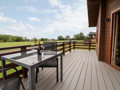 A balcony with a table two chairs a bottle and glasses overlooking a green field at Callow Lodge 15 Beaconsfield Farm Holiday Park near Shrewsbury