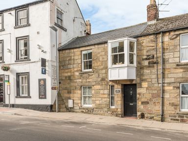 A street view showing a stone house with a black door and bay window next to a white building with window signs at Jackson Cottage in Alnmouth