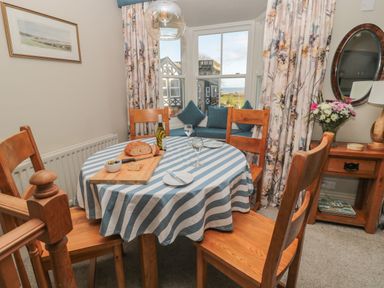 A dining area with a round table covered with a striped tablecloth four wooden chairs window seat with blue cushions bay window with floral curtains wooden side table with flowers and a mirror at Jackson Cottage in Alnmouth