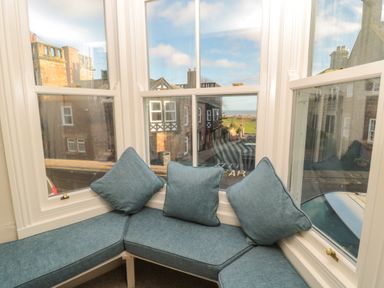 A window seat with blue cushions and pillows overlooking a street with buildings and a distant view of green fields at Jackson Cottage in Alnmouth