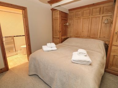 A bedroom with a bed covered with a beige bedspread and folded white towels on top next to a wooden headboard and an open door to a bathroom at Jackson Cottage in Alnmouth