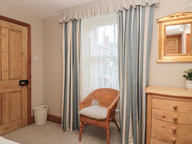 A room with a wooden door a wicker chair with a cushion a window with striped curtains a wooden dresser and a mirror at Jackson Cottage in Alnmouth