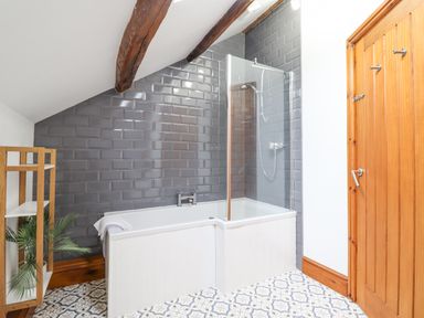 A bathroom with a bathtub and shower next to a wooden door with tiled floor and grey tiled wall at Isallt in Llanfair near Harlech